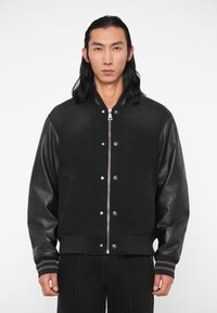 Cazadora negra tipo bomber con mangas de cuero y cuerpo texturizado, que presenta una cremallera plateada y botones a presión, acentuada con puños a rayas.