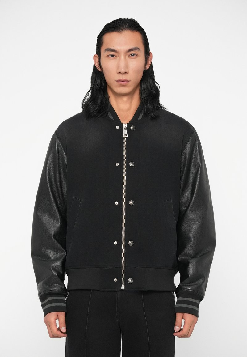 Cazadora negra tipo bomber con mangas de cuero y cuerpo texturizado, que presenta una cremallera plateada y botones a presión, acentuada con puños a rayas.