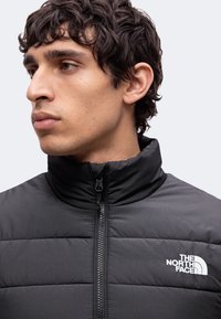 Jeune homme aux cheveux bouclés et foncés, portant une doudoune noire North Face à col montant, regardant vers la gauche.