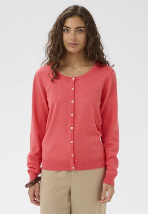 Žena s vlnitými hnedými vlasmi, oblečená v korálovom kardigáne na gombíky a béžových nohaviciach, stojaca na jednoduchom svetlom pozadí.