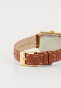 Bracelet de montre en cuir véritable marron avec coutures blanches, quincaillerie en métal couleur or et boîtier de montre carré avec un dos transparent.