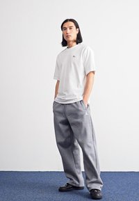 T-shirt bianco a maniche corte con un piccolo logo, abbinato a pantaloni della tuta grigi larghi e sneakers nere, il tutto su uno sfondo semplice.