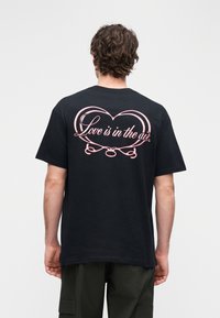 T-shirt preta com um grande coração cor-de-rosa na parte de trás, acompanhado do texto "O amor está no ar." Mangas curtas e corte descontraído.
