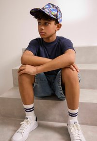 Navy színű póló, kopott farmer rövidnadrág, fehér sneaker navy kiegészítőkkel és több színben mintázott sapka. Világosszürke lépcsőkön ül.