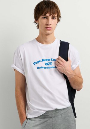 Giovane uomo con t-shirt bianca con testo blu e pantaloni grigi, che tiene la tracolla nera dello zaino sulla spalla su sfondo uniforme.