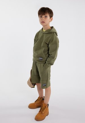 Un garçon portant un hoodie vert olive et un short, tenant une casquette beige, se tenant avec les mains dans les poches et portant des bottes en cuir marron à lacets.