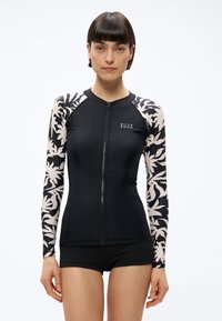 Moteris dėvi juodą užtrauktuku priekyje užsegamą apsauginę kepuraitę su baltomis gėlėtoms rankovėmis ir juodus šortus, stovinti priešais baltą foną.