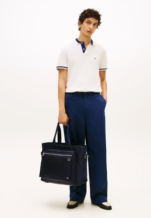 Junger Mann in weißem Polohemd und blauer Hose, der eine große schwarze Tragetasche hält und vor einem einfarbigen hellen Hintergrund steht.