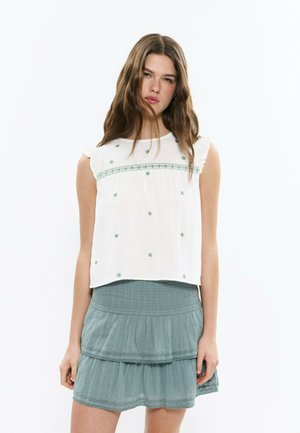 Junge Frau mit langen braunen Haaren, die ein weißes ärmelloses Oberteil mit grüner Sternstickerei und einen mehrlagigen hellgrünen Rock trägt.