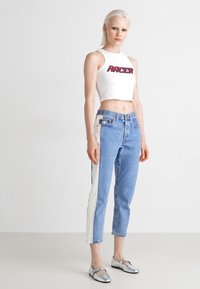 Biely crop top s nápisom "RACER" v červenom, skombinovaný s modrými džínsami s bielymi bočnými pruhmi. Model nosí strieborné baleríny.