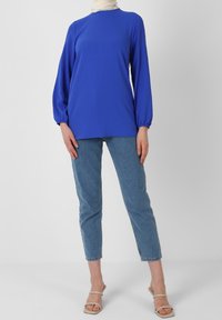 Maglia a maniche lunghe blu elettrico con collo alto e polsini elasticizzati, abbinata a jeans slim fit blu chiaro e sandali con tacco beige.