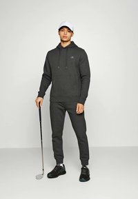 Sweat-shirt gris foncé avec capuche et poche avant, associé à un pantalon de jogging assorti. Les chaussures sont noires avec des accents bleus, et une casquette blanche complète le look.