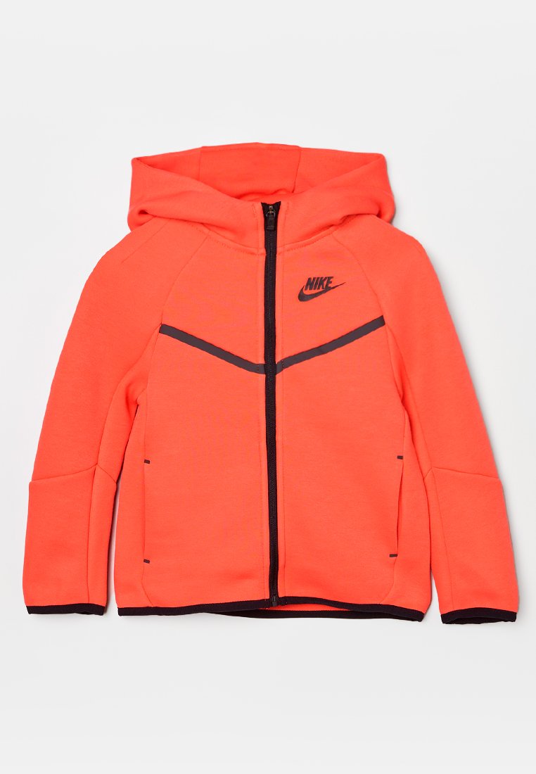 Jasnopomarańczowa kurtka z kapturem z zamkiem błyskawicznym z przodu. Posiada czarne akcenty oraz logo Nike na lewej piersi. Wykonana z gładkiego, elastycznego materiału.
