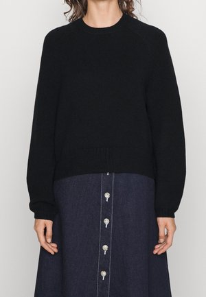 Femme portant un pull noir à manches longues et une jupe boutonnée bleu foncé avec des coutures blanches visibles, debout devant un fond uni.