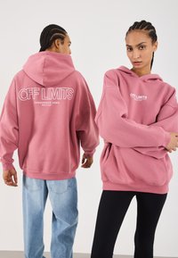 To personer med flettehår har på seg oversized dusty rose hettegensere med teksten "OFF LIMITS Dangerous Area Berlin 2001"; den ene ser fremover, mens den andre ser bakover.