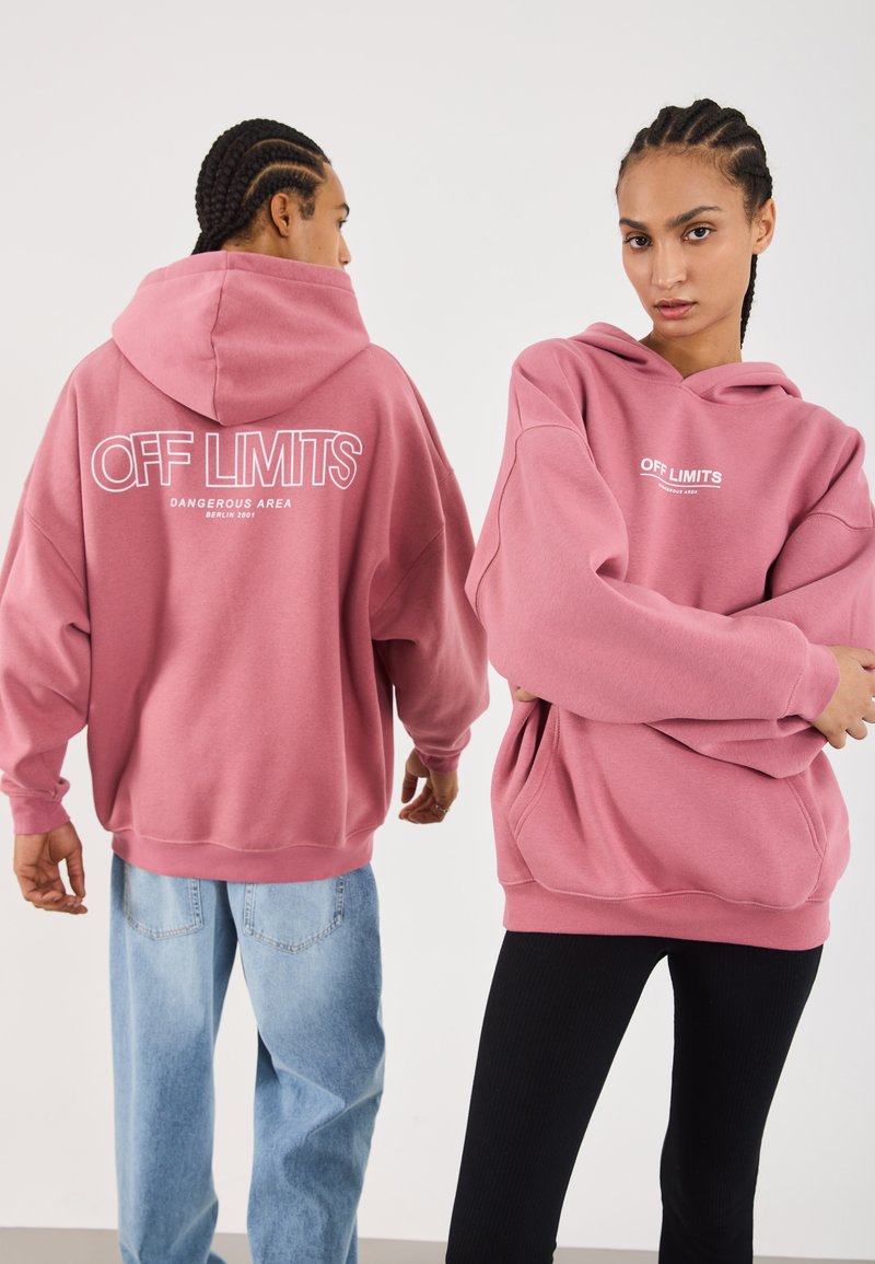 To personer med flettehår har på seg oversized dusty rose hettegensere med teksten "OFF LIMITS Dangerous Area Berlin 2001"; den ene ser fremover, mens den andre ser bakover.