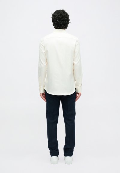 Béžová button-up košile s dlouhými rukávy, zadním prošitím, kombinovaná s tmavými kalhotami a bílými teniskami. Hladká textura, střih na míru.