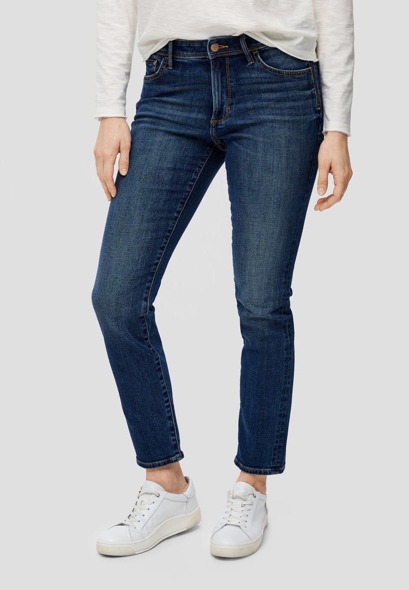 Jean en denim bleu foncé, coupe slim avec poches avant, passants de ceinture et fermeture à bouton. Associés à des baskets blanches à lacets.
