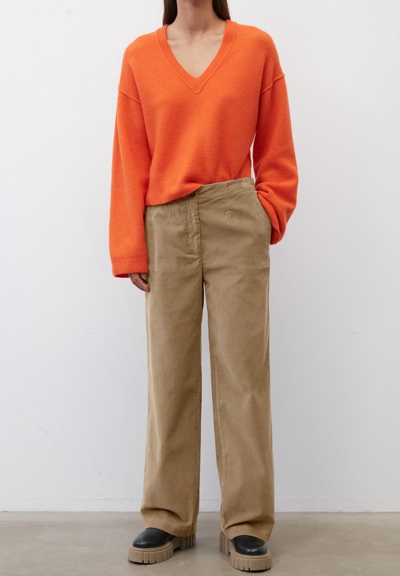 Personne portant un pull orange vif à col en V, un pantalon beige ample et des chaussures noires à semelle épaisse, debout devant un mur blanc uni.