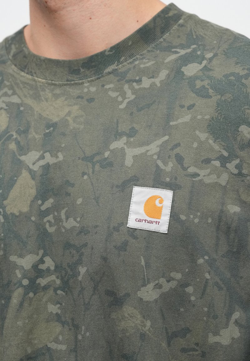 Close-up of a person wearing a green and gray camouflage-patterned shirt with a small white square Carhartt logo patch on the chest.