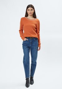 Oranje gebreide trui met een gestructureerde afwerking en versierde halslijn, gecombineerd met blauwe spijkerbroek en zwarte enkellaarsjes.