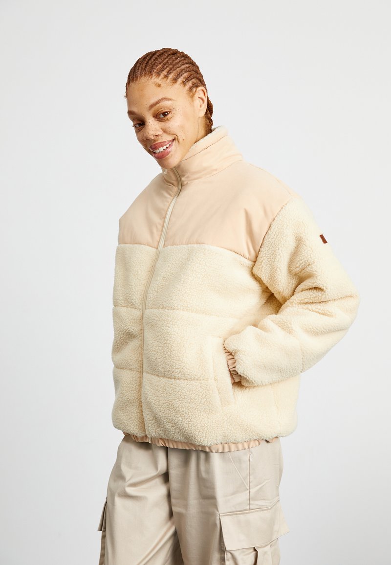 Fleecejacka i off-white med en beige slät tygöppning, dragkedjestängning och elastiska muddar. Har en boxig passform och sidfickor.