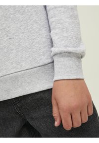 Felpa grigia con superficie strutturata e polsini a coste, abbinata a jeans in denim scuro, con una mano appoggiata sulla coscia.