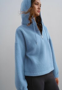 Lysblå fleece pullover med lynlås i halsen, stor hætte og vide ærmer, med en blød, struktureret overflade.