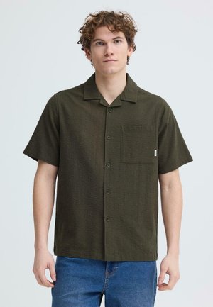 Jeune homme aux cheveux bouclés portant une chemise boutonnée manches courtes vert foncé avec une poche poitrine et un jean bleu, sur fond uni.