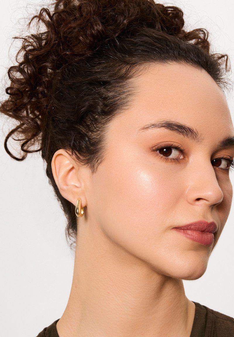 Złoty kolczyk w kształcie obręczy o gładkiej, połyskującej powierzchni. Zakrzywiony design, średni rozmiar, noszony na małżowinie ucha, uzupełniający neutralny strój.