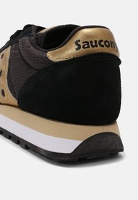 Talons de chaussure de sport noirs et dorés avec le logo "Saucony" sur un panneau métallique doré, une semelle intermédiaire beige et une semelle extérieure noire à crampons.