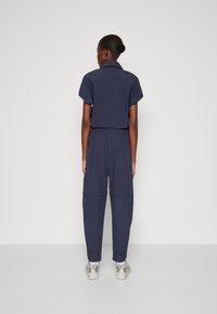 Navy jumpsuit met korte mouwen en elastische taille. Voorzien van ritssluitingen en taps toelopende pijpen. Materiaal lijkt lichtgewicht met een minimale textuur.