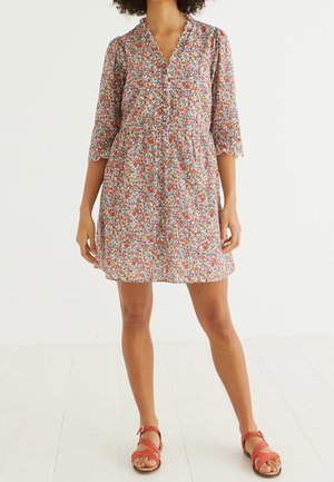 Kvinde iført en kort, blomsterprintet kjole med bølgede kanter og trekvartlange ærmer, kombineret med røde åbne sandaler.