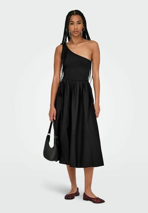 Abito nero a spalla unica con corsetto aderente e gonna svasata a pieghe. Il modello indossa delle scarpe ballerine rosse con pois bianchi e tiene una borsa nera.