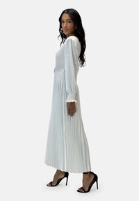 Vestido blanco plisado con mangas largas, corpiño entallado y puños fruncidos. Combinado con sandalias negras de tacón alto. Diseño sencillo y elegante.