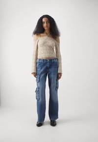 Béžový háčkovaný top s off-shoulder dizajnom a dlhými rukávmi, skombinovaný s voľnými modrými denimovými cargo nohavicami so bočnými vreckami.
