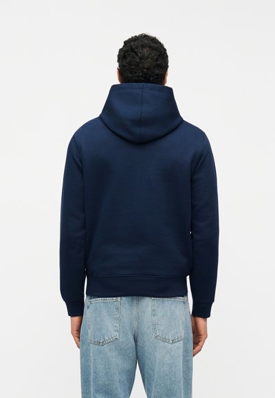 Homme portant un sweat à capuche bleu marine et un jean en denim bleu clair, vu de dos sur un fond blanc uni.