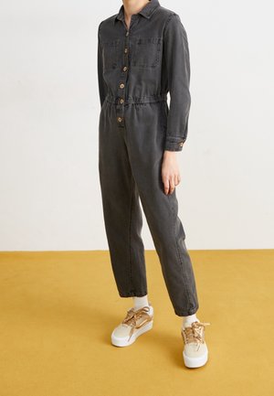 Osoba ubrana w wyblakły czarny kombinezon na guziki z elastycznym pasem i beżowe trampki, stojąca na podłodze w kolorze musztardowej żółci.