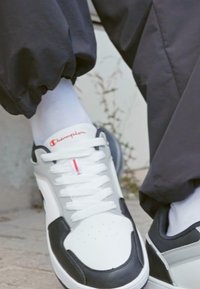 Adidași albi și gri cu accente negre, având un bot perforat, șireturi albe și un logo roșu pe limbă.
