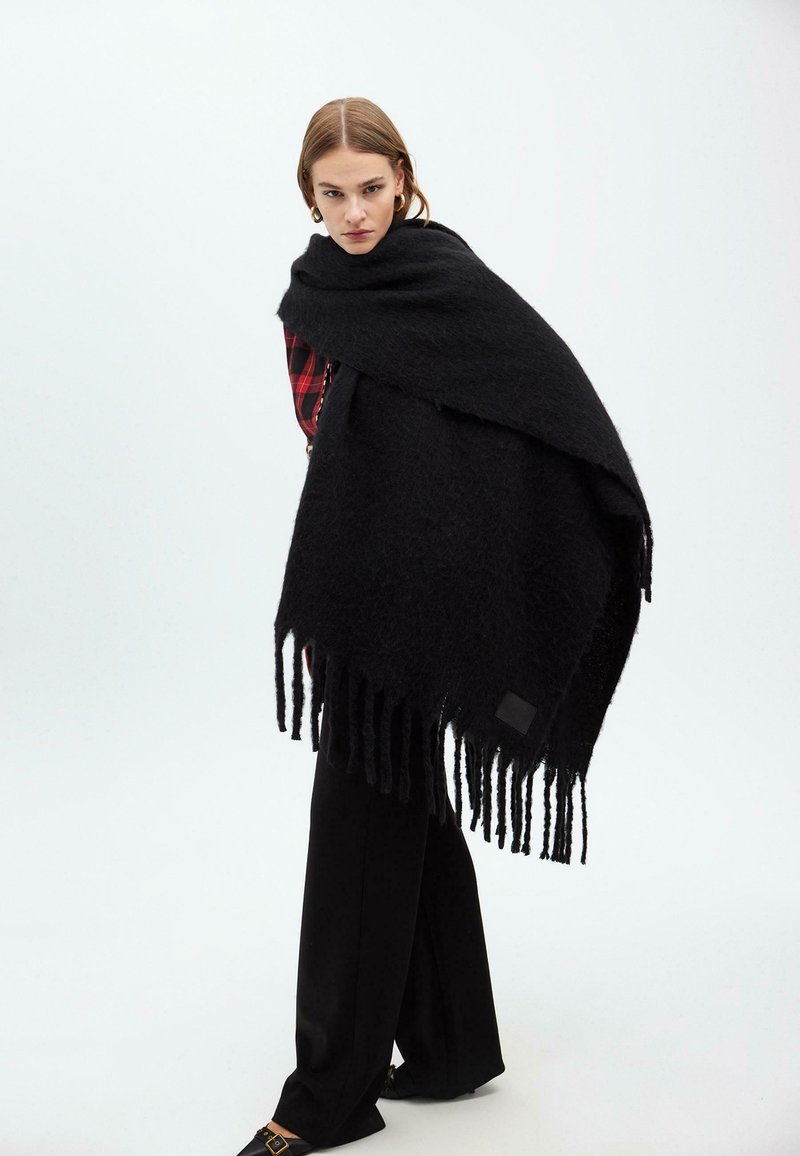 Černý pletený šál s třásněmi, vyrobený z texturované látky. Modelka ho má přehozený přes ramena a kombinuje ho s červenou kostkovanou blůzou.