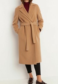 Beige, lång kappa i ull-blandning med sjalkrage och bälte i midjan. Har sidofickor och en rak silhuett, ihop med svarta byxor.