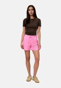 Γυναίκα που στέκεται φορώντας ένα εφαρμοστό σκούρο καφέ κοντομάνικο τοπ, ροζ σορτς με κορδόνι και μπεζ αθλητικά παπούτσια μπροστά σε απλό φόντο.