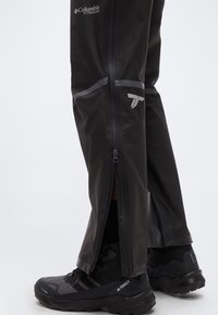 Μαύροι αδιάβροχοι παντελόνες με πλαϊνές φερμουάρ, φορεμένοι πάνω από μαύρα παπούτσια πεζοπορίας Terrex, σε ανοιχτό φόντο.