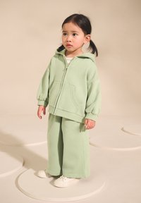 Ljusgrön sweatshirt med dragkedja och framficka, kombinerad med vida byxor och vita sneakers, mot en neutral bakgrund.