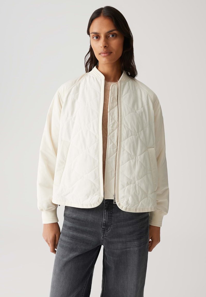 Veste matelassée blanche avec col montant et fermeture éclair, dotée d'une coupe décontractée et de manches longues, associée à un jean gris foncé.
