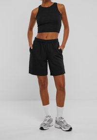 Personne portant un haut court noir sans manches et un short noir à taille élastique, debout les mains dans les poches, chaussettes blanches et baskets grises.