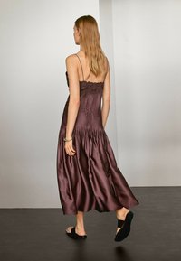 Langes, ärmelloses, dunkelburgunderfarbenes Kleid mit smockiertem Oberteil und Spitzenverzierungen, fließende Satin-Textur, kombiniert mit schwarzen Slippern.