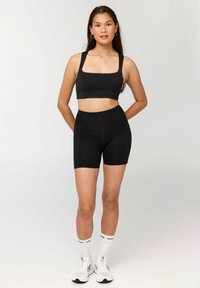 Noor naine seisab käed selja taga, seljas must sportrinnahoidja, mustad kõrge vöökohaga rattapüksid, valged sokid ja valged tossud.