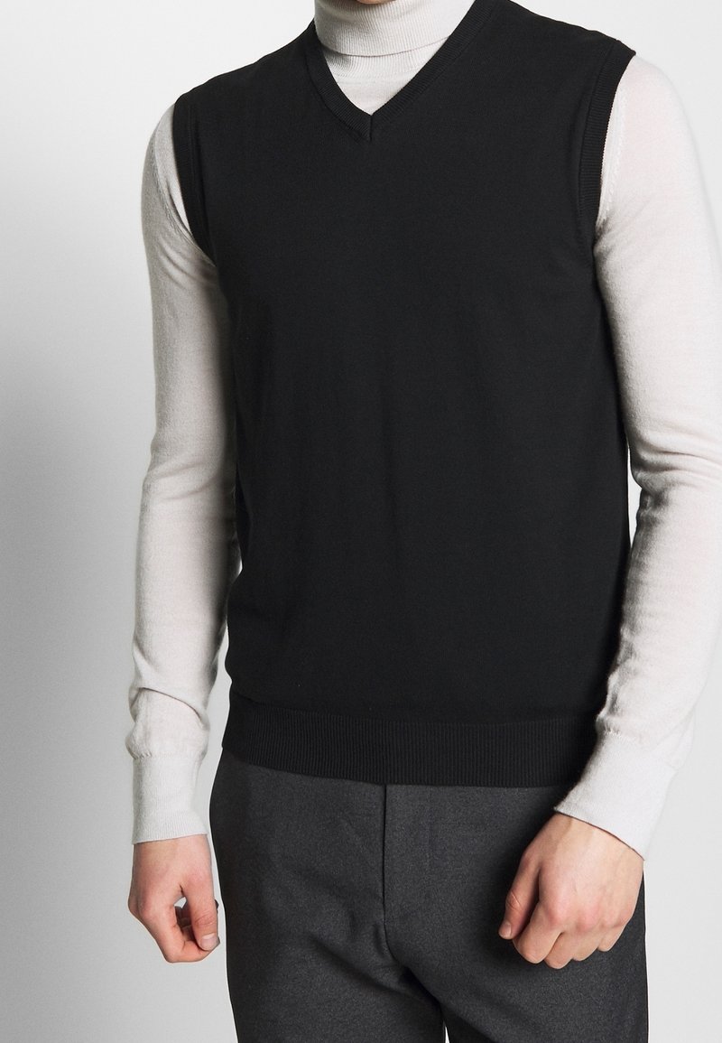 Black sleeveless sweater with a V-neck, made of soft knit material. Worn over a light turtleneck, featuring a ribbed hem.