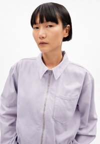 Chaqueta con cremallera en lavanda claro, con cuello, que presenta un bolsillo en el pecho, material de algodón suave y un corte relajado. Textura lisa y sutiles detalles en las costuras.